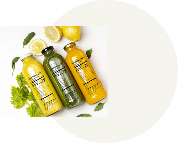 A white circle is displayed on a grey background with a square showing green, yellow and orange juices in glass bottles lying next to each other and greenery behind that.