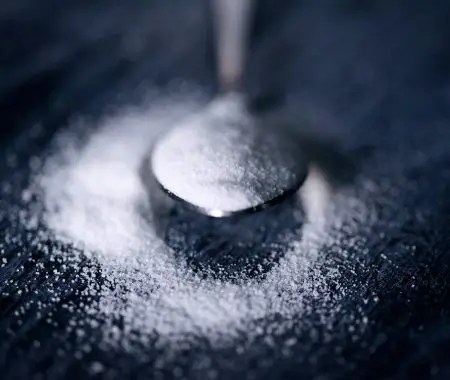 A spoon full of sugar sitting on a black table top with some sugar sprinkled around it