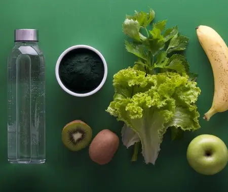 Bottle of water, green powder, lettuce, parsley, kiwi, banana and apple on green table