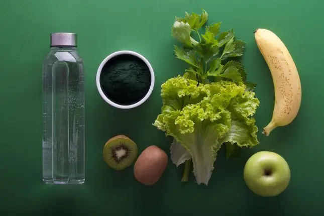 Bottle of water, green powder, lettuce, parsley, kiwi, banana and apple on green table