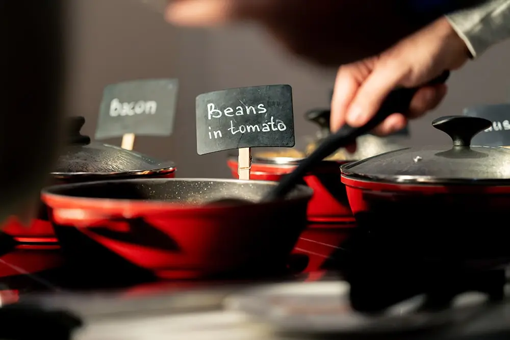 A person dishing up food from a pan with a clear buffet label that reads "Beans in tomato"