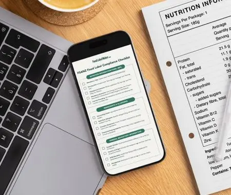 Workspace showing laptop, smartphone displaying FSANZ food label compliance checklist, printed nutrition information panel, and coffee cup on wooden desk