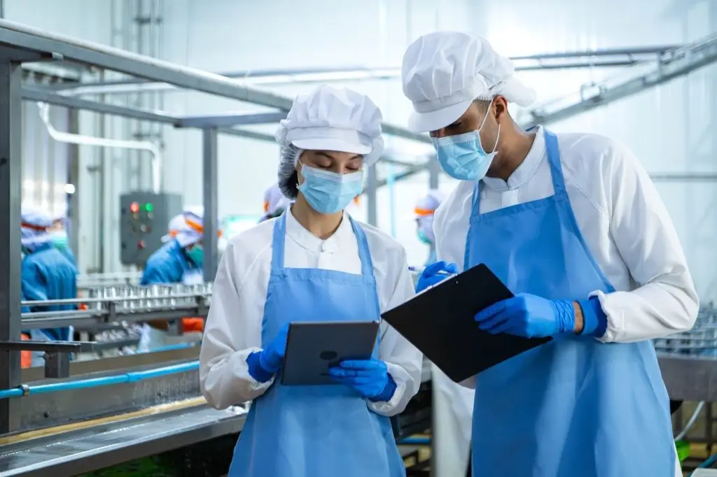 Two quality control workers in protective clothing and face masks are analysing data on a tablet and clipboard in front of a food production line.