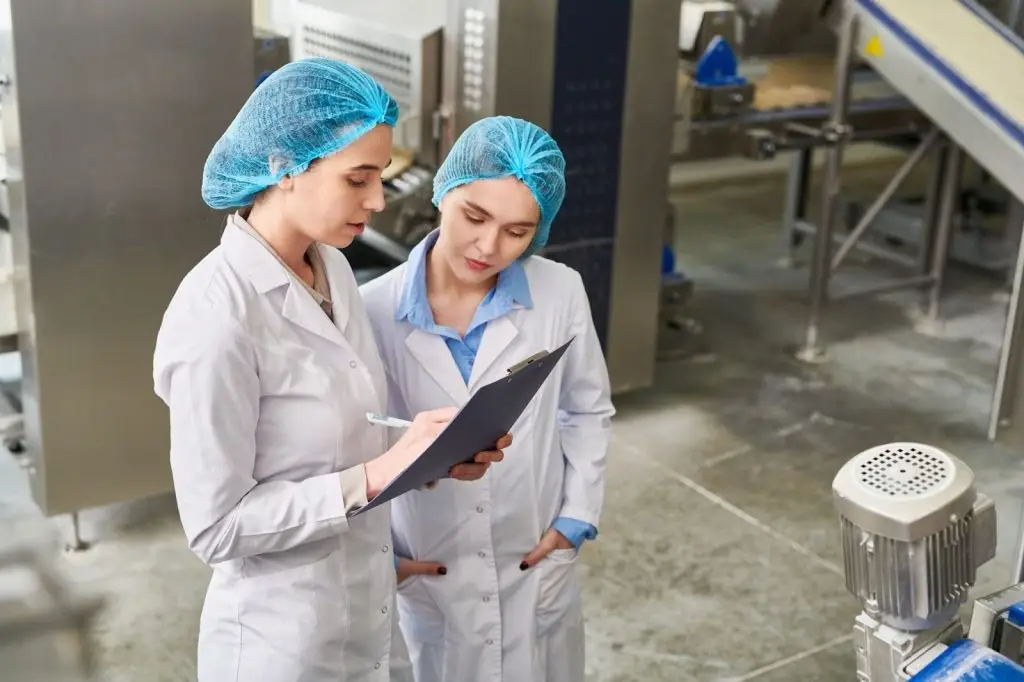 Two food production workers in lab coats and hair nets are reviewing a checklist on a clipboard inside a factory.