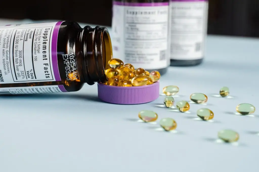 An open brown supplement bottle tipped over, spilling yellow softgel capsules into its purple lid.